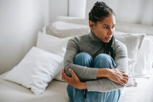 Black woman sitting on bed clasping her knees experiencing anxiety about anxiety