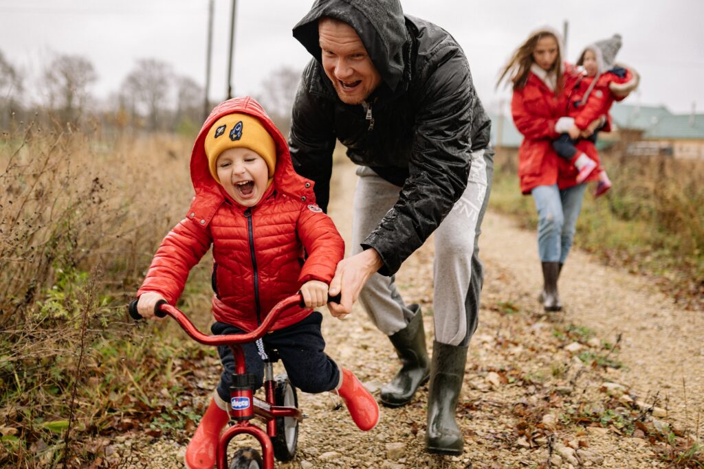 A young family on the road to successful parenting with the dad training his young son how to ride a bike and the mom walking behind them.